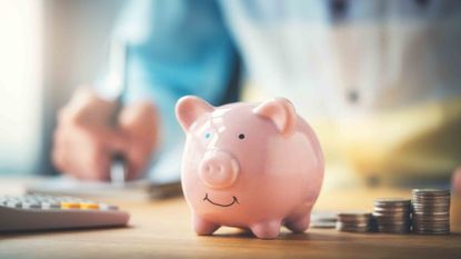 A person sitting at a desk, holding a pen, with a calculator, piggy bank and a stack of coins