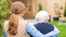 A woman puts her arm around an older man walking outside. 