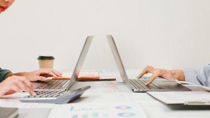 Back-to-back laptops on a desk with only the hands of people working on them showing.