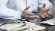 Doctor gestures with his hands above desk that holds files and a stethoscope.