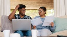 A couple on their couch with a laptop look over bills while he scratches his head.