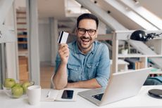 A happy young man in a modern apartment smiles, holding a credit card with his laptop open.