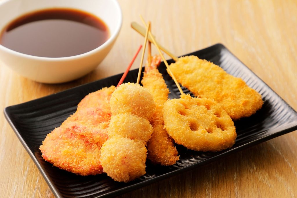 Kushikatsu (deep-fried skewered seafood, meat, and vegetables) on a plate with a bowl of sauce on the side for dipping.