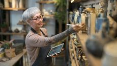 An older woman holding a tablet works in a pottery shop.